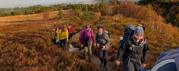 A group of women are walking uphill along a narrow dirt path through autumnal moorland. The women are wearing outdoor clothing and carrying backpacks of various sizes. The woman closest to the camera on the right has a large backpack with gear attached and is wearing a patterned head covering. The women behind are dressed in colourful jackets, including shades of green, yellow, purple and grey. The path winds between dense heather and low shrubs in reddish‑brown and golden tones.